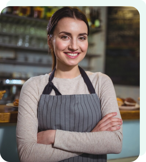 A smiling woman in an apron stands confidently in front of a well-stocked bar, exuding warmth and hospitality.